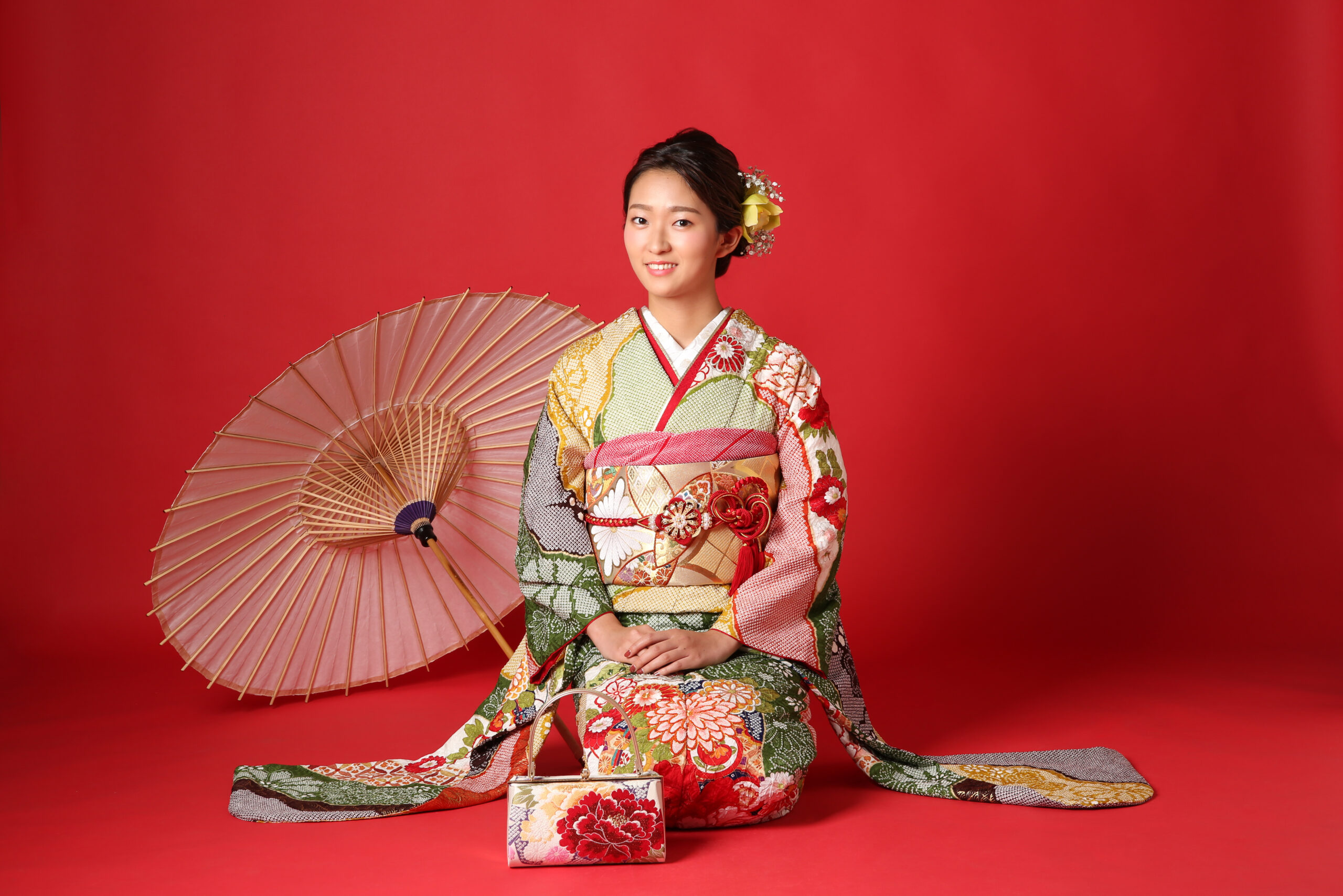 赤背景のスタジオで撮影された総絞り・辻が花振袖。赤を基調に緑や金彩の花柄が映える成人式向け振袖で、和傘と草履バッグを合わせた前撮り全身写真。